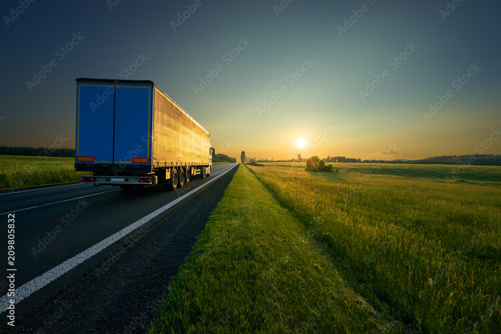 Obraz premium Truck driving on the asphalt road in a rural landscape in golden sunset colors