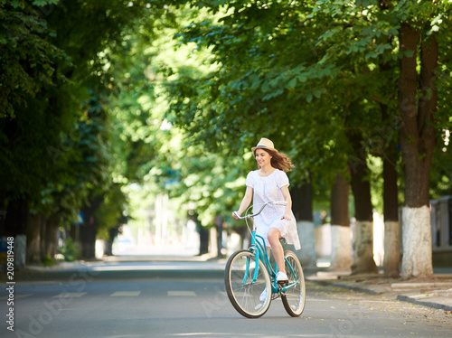 Concentration. Smiling female in white dress and straw hat riding blue bicycle not looking at the camera on green wide street with trees around. Beautiful girl excitement park happiness joy summer