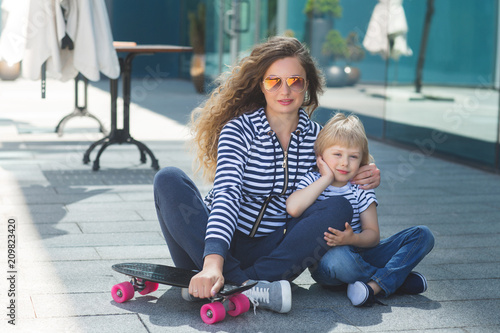 Young beautiful mother with her little cheerful son having fun and skating outdoors. Pretty cute kid with his mom together resting outside with skateboard. Happy family