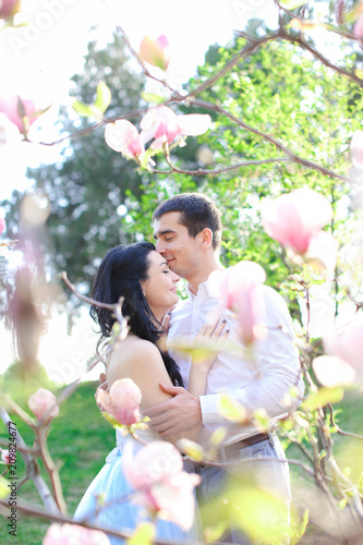 Brunette boy hugging and kissing caucasian girl near magnolia blooming tree. Concept of love and spring season.