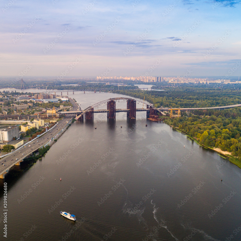 Panoramic view of a modern city with a river, unfinished bridge and park part of the city. Skyline bird eye aerial view with industrial zone and high-rise residential area under dramatic cloud sunset