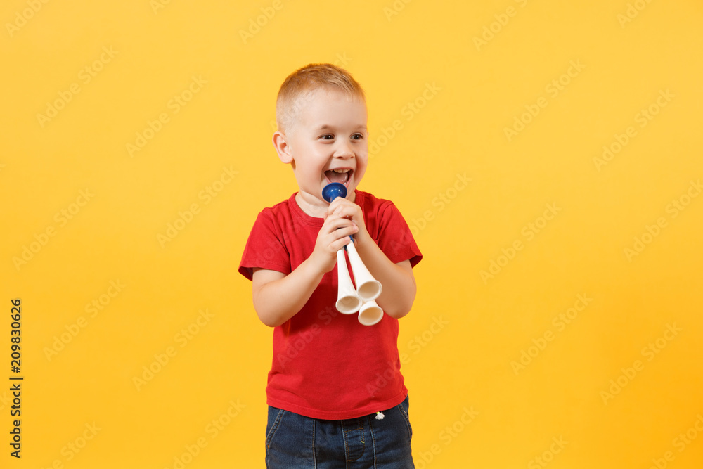 Little cute kid baby boy 3-4 years old, football fan in red t-shirt holding in hand pipe, blowing isolated on yellow background. Kids sport family leisure lifestyle concept. Copy space advertisement. Little cute kid baby boy 3-4 years old, football fan in red t-shirt holding in hand pipe, blowing isolated on yellow background. Kids sport family leisure lifestyle concept. Copy space advertisement.