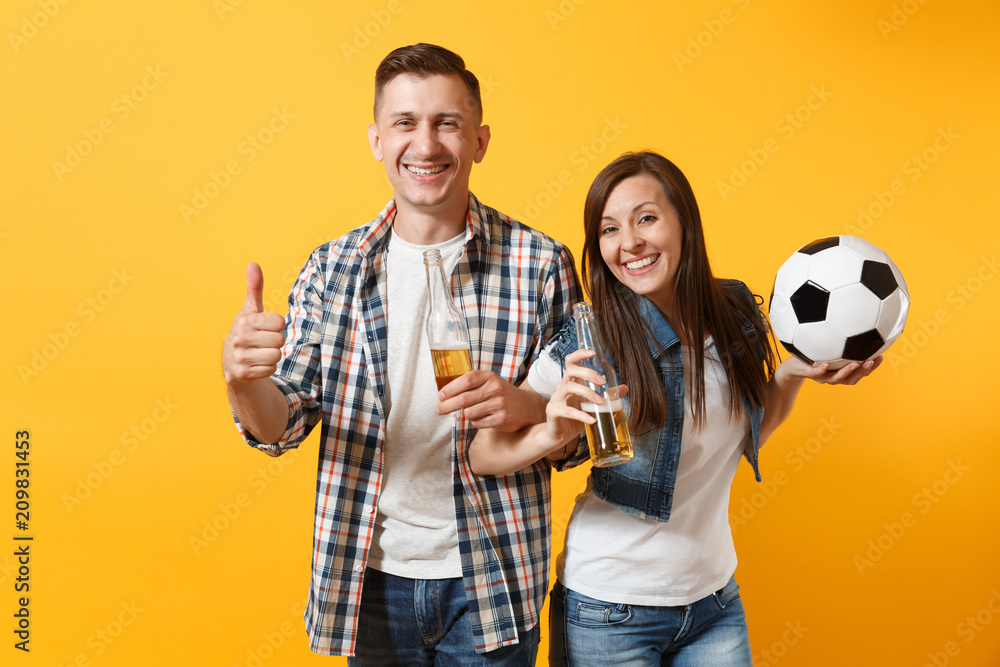 Young happy cheerful couple supporter, woman man, football fans cheer up support team, holding beer bottle, soccer ball isolated on yellow background. Sport, family leisure, people lifestyle concept. Young happy cheerful couple supporter, woman man, football fans cheer up support team, holding beer bottle, soccer ball isolated on yellow background. Sport, family leisure, people lifestyle concept.