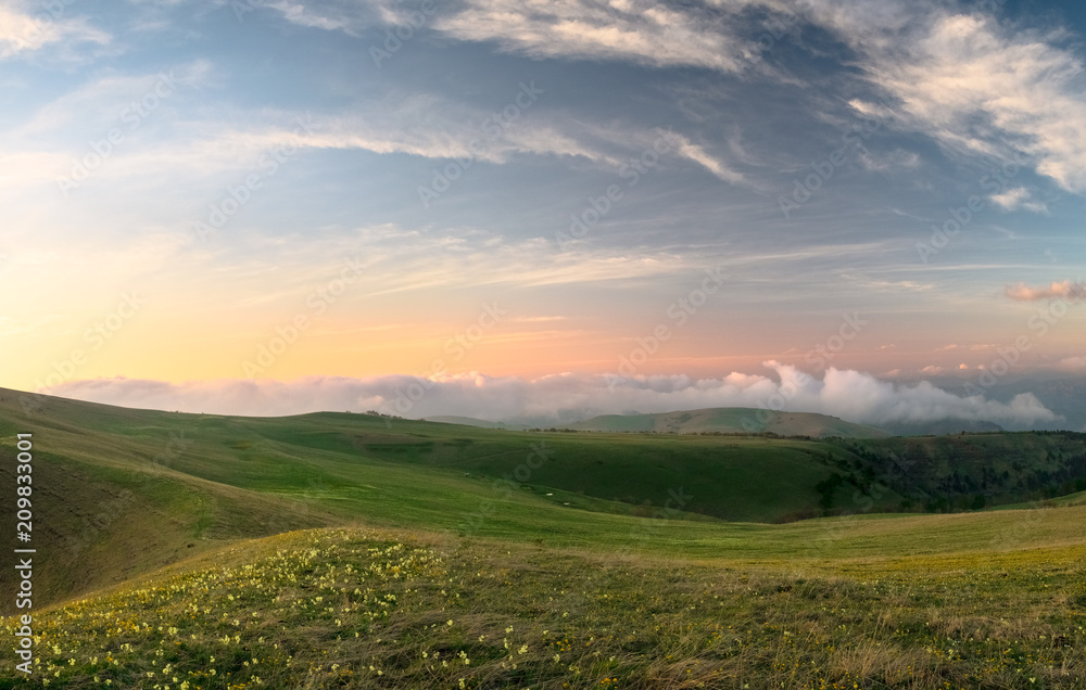 Alpine meadows of the Karachay-Cherkess Republic with green grass and flowers with a colored sky and clouds in the evening sunset in the spring