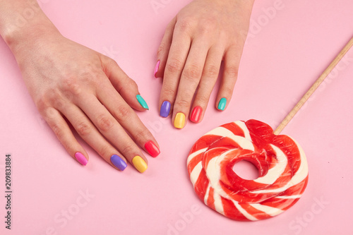 Female polished nails and lollipop. Woman hands with varnished nails and big lollypop on colored table, top view. Candy colored nails.