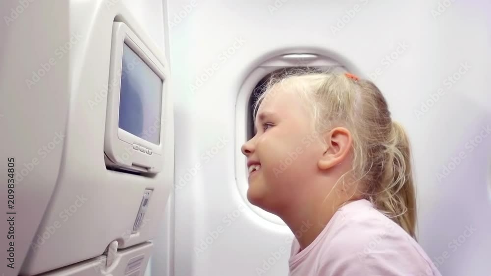 A little girl traveling in an airplane, sitting in her seat, playing with a built-in video player and choosing a cartoon watched by a parent A little girl traveling in an airplane, sitting in her seat, playing with a built-in video player and choosing a cartoon watched by a parent