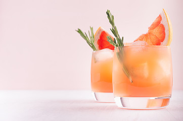 Two elegance cold wet drinking glasses with fresh grapefruit summer cocktail, ice, rosemary on pink background.