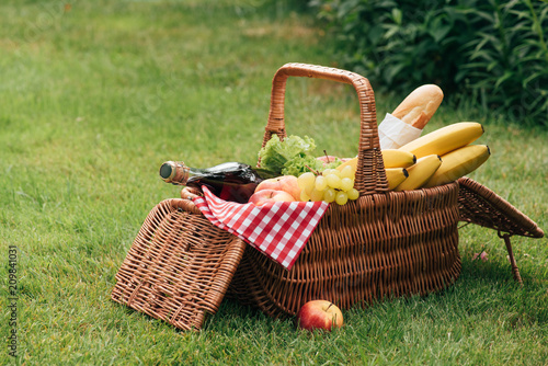 tasty fruits and bottle of champagne in wicker basket on green grass at picnic