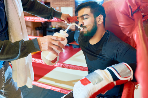 Crop senior coach helping young bearded boxer in gloves with drinking water while sitting in corner of ring. 