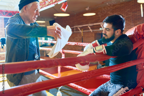 Bearded man sitting in ring corner while elderly trainer waving with towel for refreshment. 