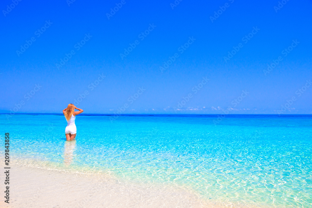 Gorgeous blond woman with white clothes is standing in the water at the sandy beach