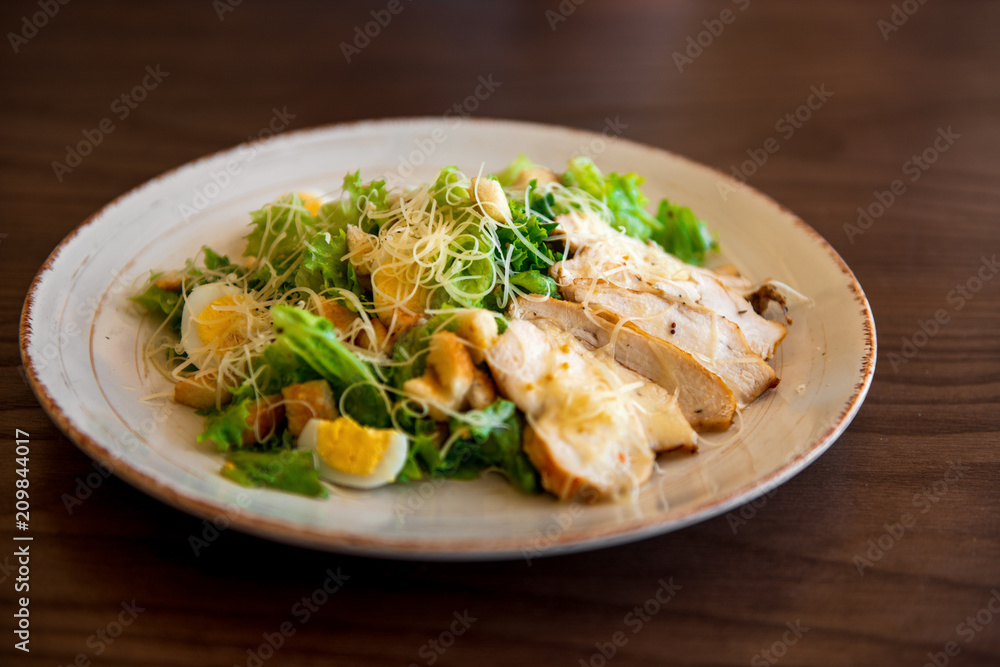 Fresh summer salad Ceasar with eggs, lettuce, chicken and parmesan cheese on white plate on wooden background; healthy concept; side view