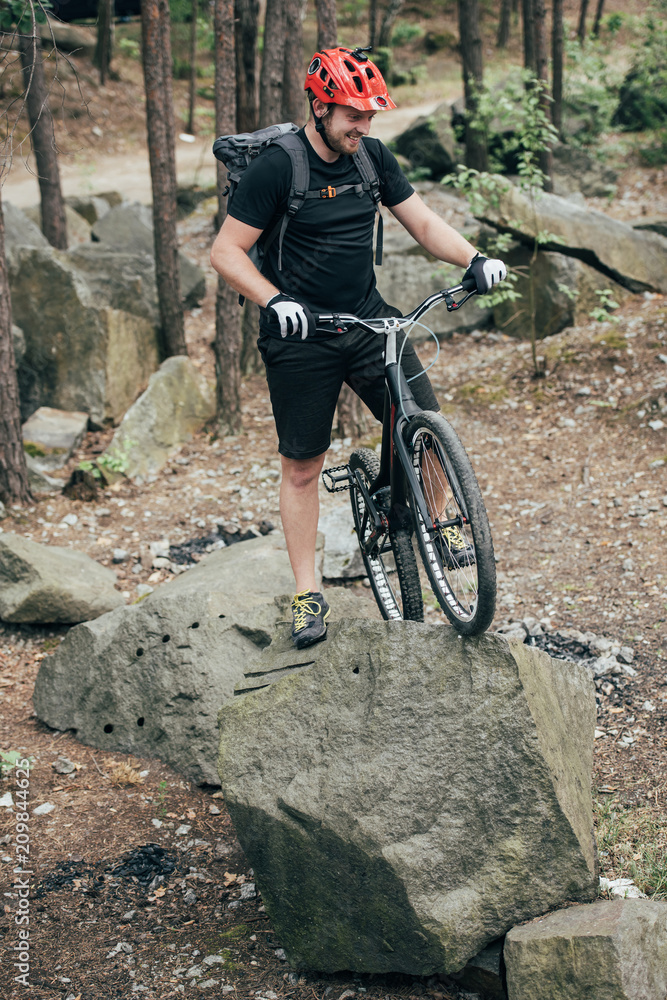 Fototapeta premium smiling male extreme cyclist in protective helmet standing with mountain bicycle on stone in forest