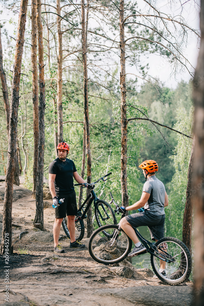 Fototapeta premium Two male extreme cyclists in helmets with mountain bikes resting with sport bottles of water in forest