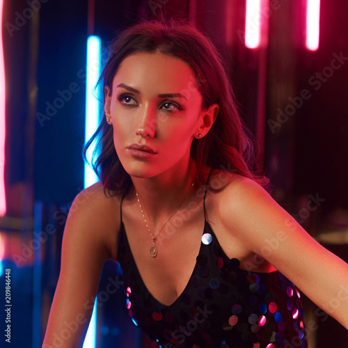 Brunette woman wearing elegant black sequin strap cami dress sitting on large cube with crossed legs. Attractive caucasian female model posing against glowing neon lamps and mirrors on background