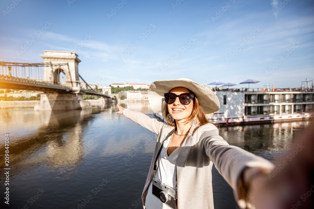 Happy young woman tourist making selfie photo standing in front of the old bridge in Budapest city