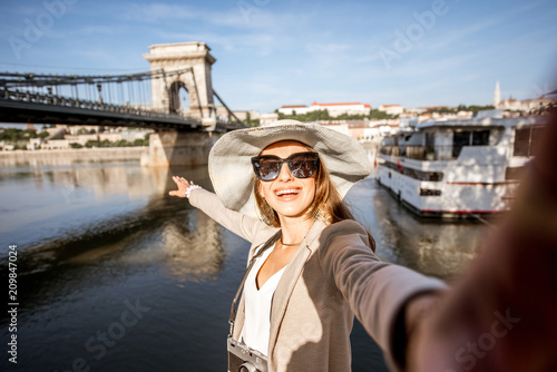 Happy young woman tourist making selfie photo standing in front of the old bridge in Budapest city