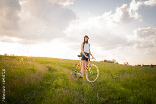 Girl with bicycle in beautiful rural area. Young pretty female person with retro bike standing in a meadow on bright sunny afternoon in summer