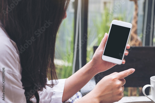 Mockup image of a woman holding and showing white mobile phone with blank black screen in cafe