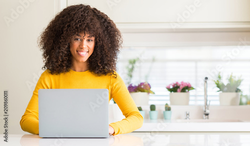 African american woman using computer laptop at kitchen with a happy face standing and smiling with a confident smile showing teeth