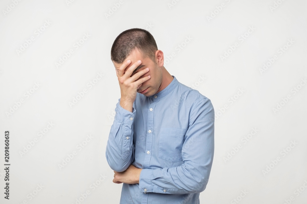 Displeased young man in blue shirt covering his eyes over gray background Displeased young man in blue shirt covering his eyes over gray background