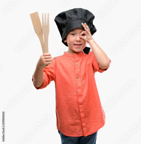 Dark haired little child wearing chef uniform with happy face smiling doing ok sign with hand on eye looking through fingers