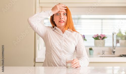 Thirsty redhead woman and glass of water stressed with hand on head, shocked with shame and surprise face, angry and frustrated. Fear and upset for mistake.