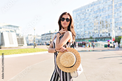 Smiling stylish  girl with dark hair breathes a full breast and enjoys summer, standing on sunny square. Portrait of adorable young woman in stripped dress having fun outside