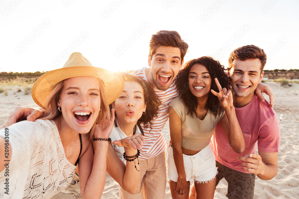 Fototapeta premium Grupa szczęśliwych młodych przyjaciół w letnie ubrania, biorąc selfie, stojąc na plaży podczas wakacji na łonie natury