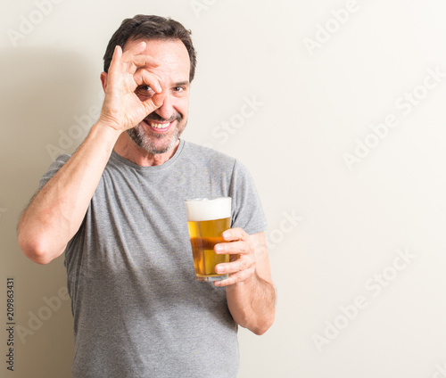 Senior man drinking beer with happy face smiling doing ok sign with hand on eye looking through fingers