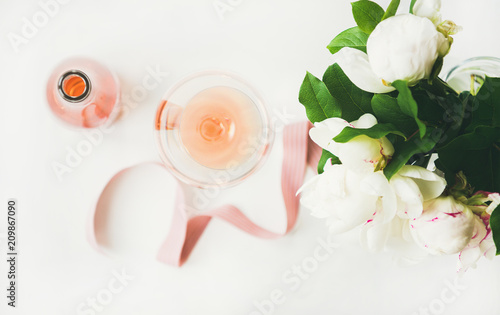 Flat-lay of rose wine in glass and bottle, pink decorative ribbon, peony flowers over white background, top view, horizontal composition. Summer celebration, wedding greeting card, invitation concept