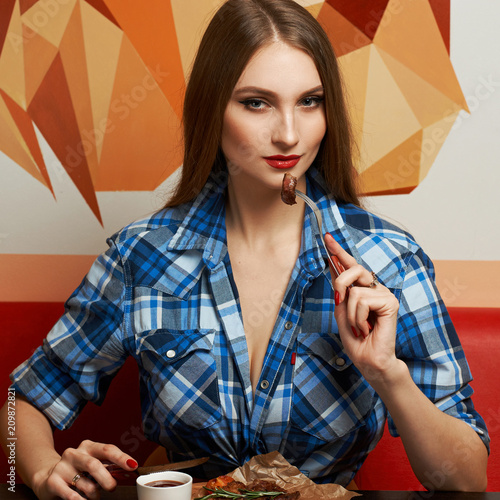 Beautiful sexy female model dressed in blue unbuttoned shirt sitting at table and biting piece of tasty grilled steak impaled on fork. Young pretty long haired woman at meat restaurant or steakhouse.