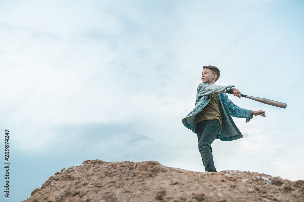 Obraz premium low angle view of boy in denim jacket playing with baseball bat while standing on hill against cloudy sky