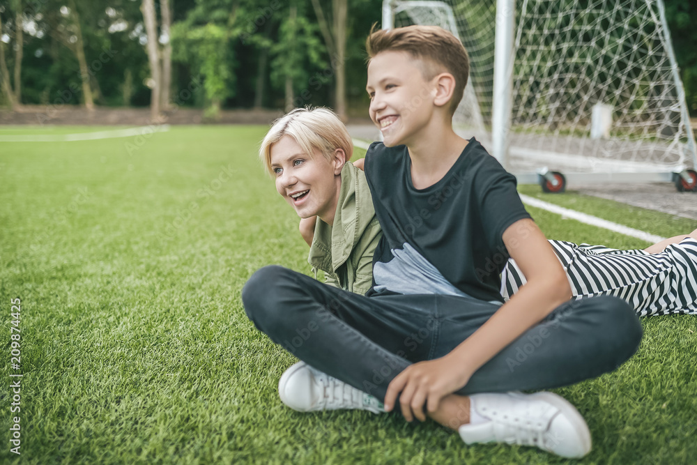 cheerful mother and son laughing and looking away while resting together on green lawn