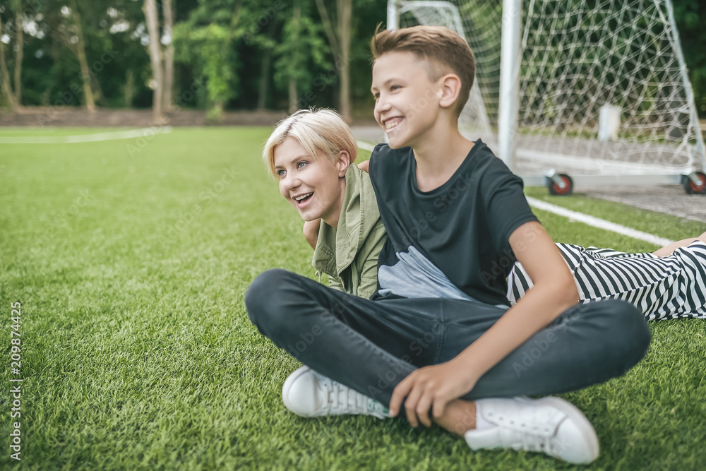 cheerful mother and son laughing and looking away while resting together on green lawn cheerful mother and son laughing and looking away while resting together on green lawn