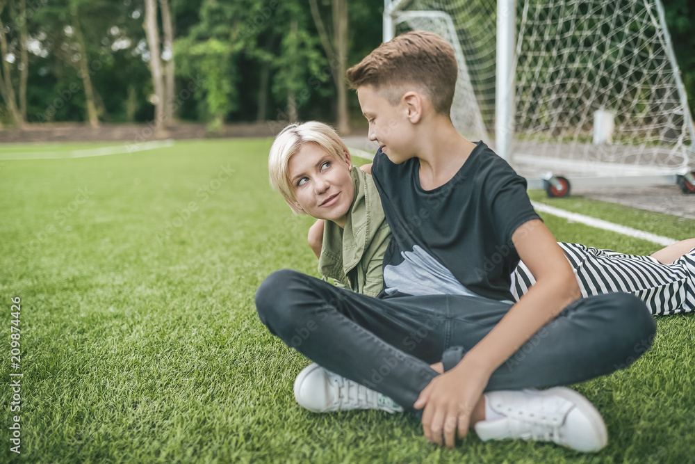 happy mother and son smiling each other while resting together on green lawn happy mother and son smiling each other while resting together on green lawn