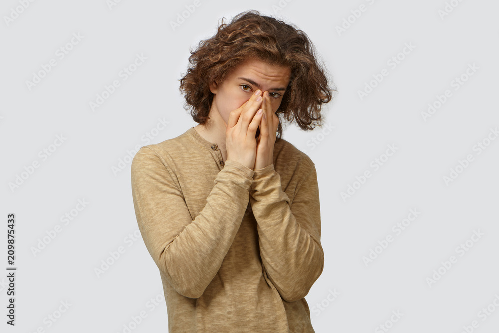 Portrait of attractive stylish displeased young man with voluminous hair covering mouth and nose with both hands, holding breath because of bad smell, looking at camera, posing isolated in studio Portrait of attractive stylish displeased young man with voluminous hair covering mouth and nose with both hands, holding breath because of bad smell, looking at camera, posing isolated in studio
