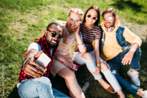 selective focus of multiethnic friends taking selfie on smartphone while resting on green lawn in park