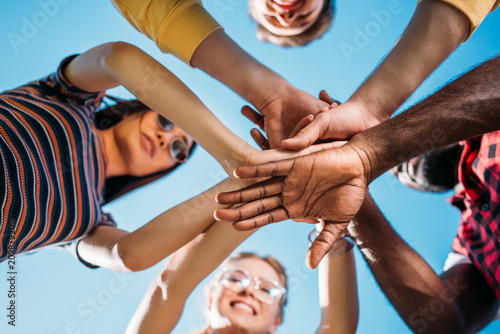 bottom view of multiracial young friends holding hands together with blue sky on backdrop