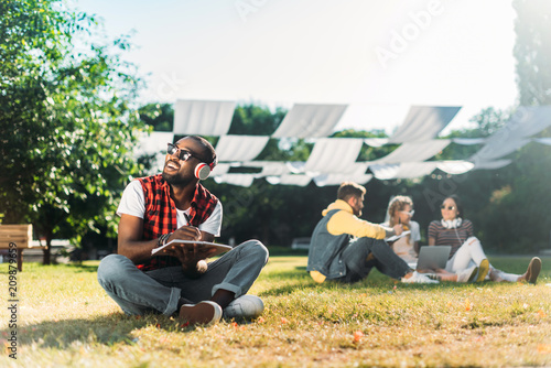 selective focus of african american man in headphones with notebook resting on green grass with multiracial friends with laptop behind