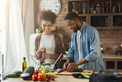 Obraz na plátně Laughing black couple preparing salad in kitchen