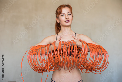 Girl with red hair dressed in a black swimsuit, holding a thick, long orange electric wire. Gray concrete walls. photo