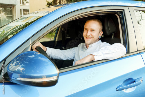 Young handsome businessman sitting behind steering wheel, test driving his new caribbean blue car. Smiling man in vehicle, satisfied with newely purchased automobile. Portrait, close up, background photo