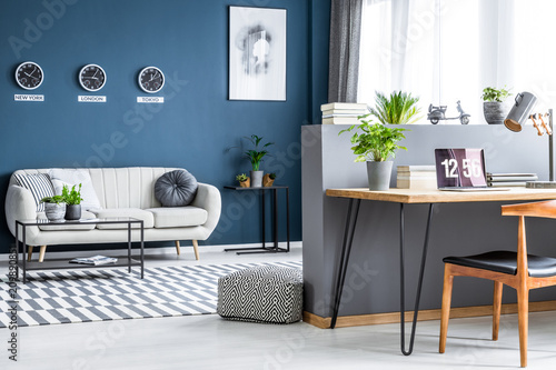 Dark blue living room interior with three clocks, simple poster, bright sofa and home office corner with laptop on hairpin desk photo