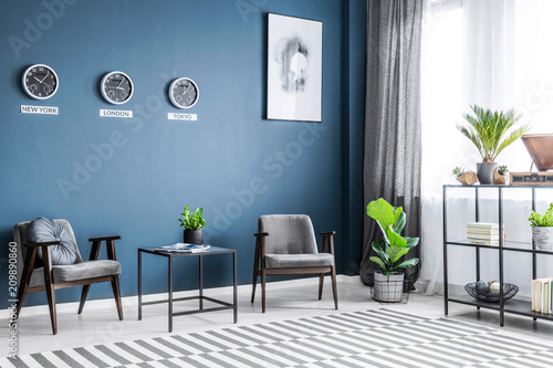 Two grey armchairs, metal rack with decor, window with drapes and fresh plants placed in dark living room interior