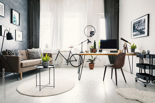 Black chair at wooden desk with computer desktop in workspace interior with sofa and bike. Real photo © Photographee.eu