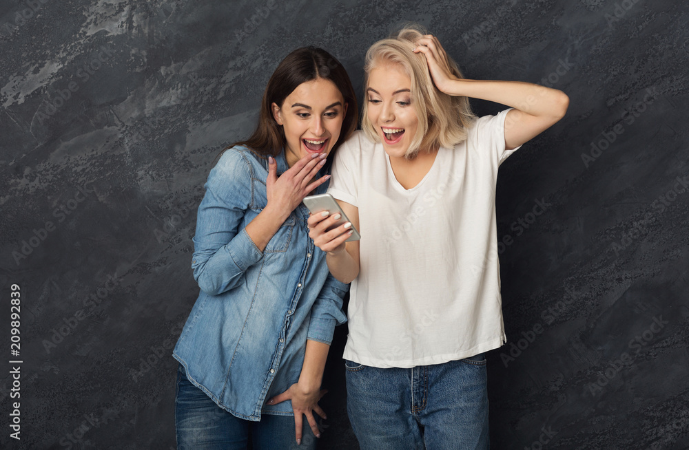 Surprised female friends using smartphone at dark studio backgro