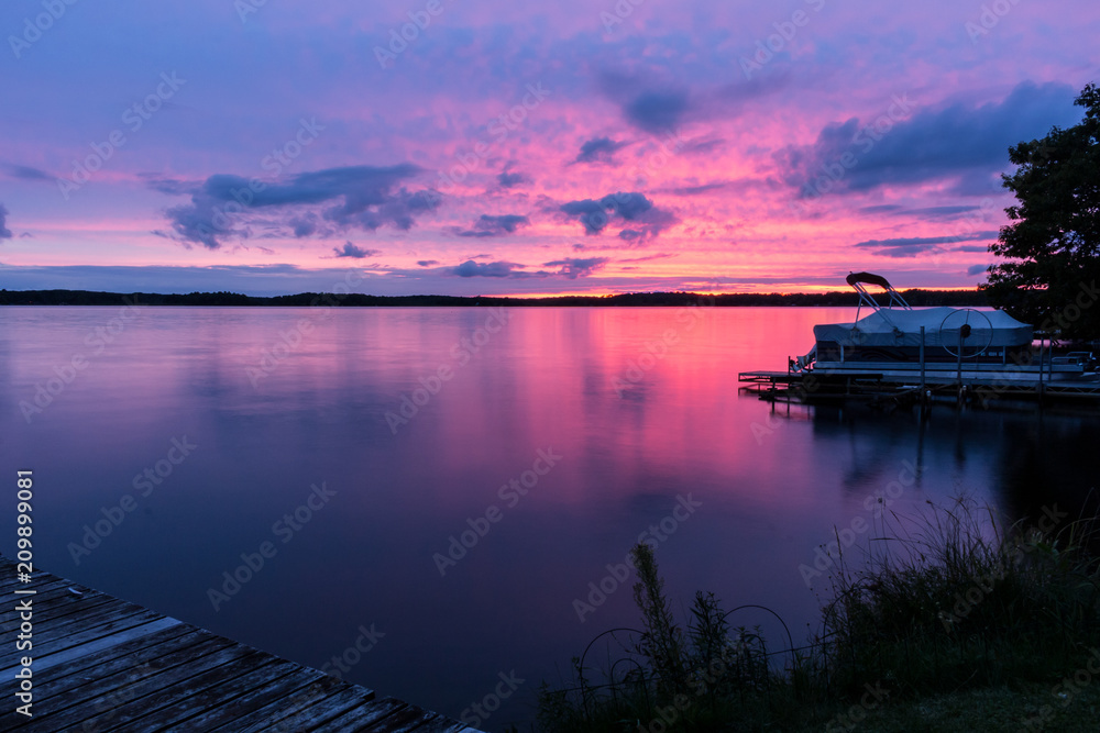 Colorful pink, purple & blue sunset over a calm lake with a pontoon boat at the shore. Beautiful northwoods scene with a colorful sky reflected in the water. Concepts of vacation, nature, travel