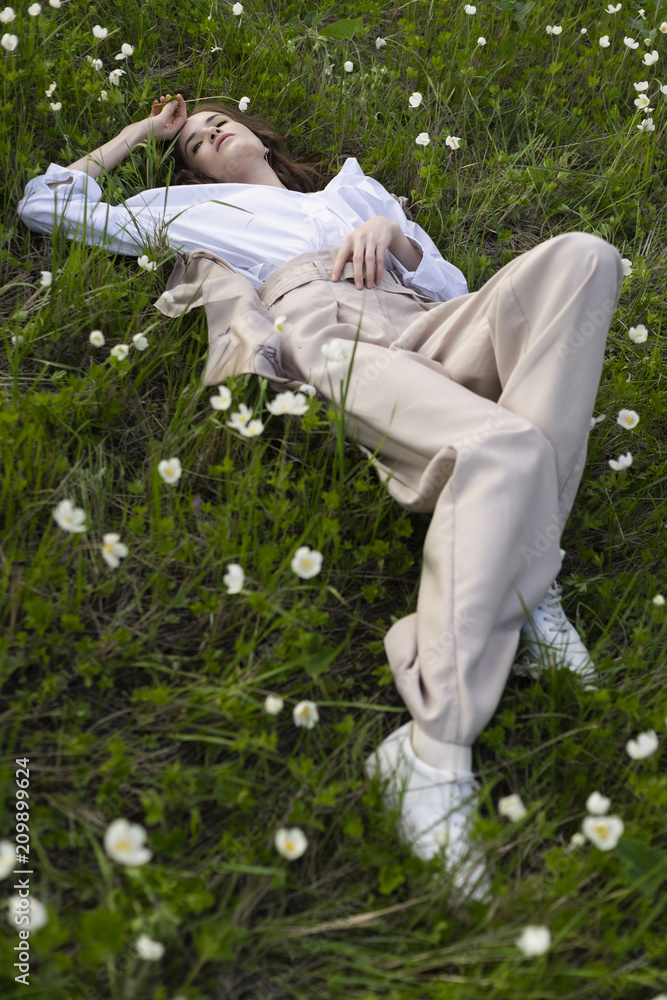 Beautiful stylish young girl wearing a trendy white shirt, beige bloomers, and a pareo lies relaxing on the green grass among the wild flowers Beautiful stylish young girl wearing a trendy white shirt, beige bloomers, and a pareo lies relaxing on the green grass among the wild flowers