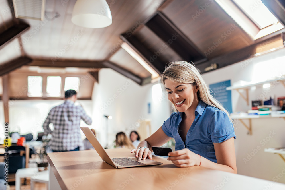 Young woman holding credit card and using laptop, coworkers in the background.